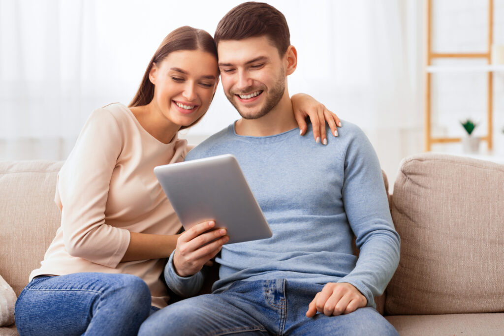 Spouses Using Digital Tablet Sitting On Couch At Home