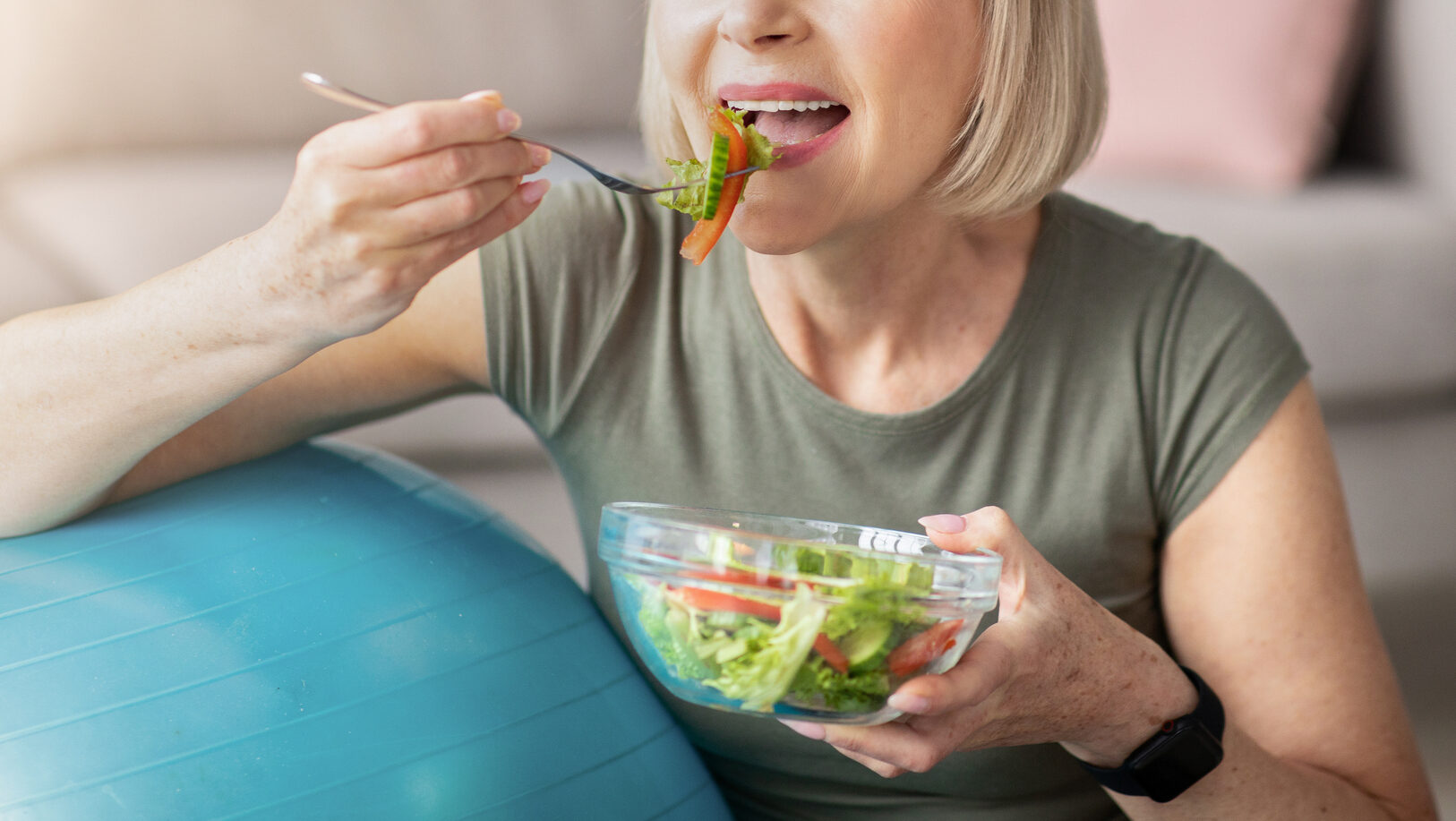 Sports and nutrition concept. Smiling senior lady leaning on fitness ball, eating fresh vegetable salad at home